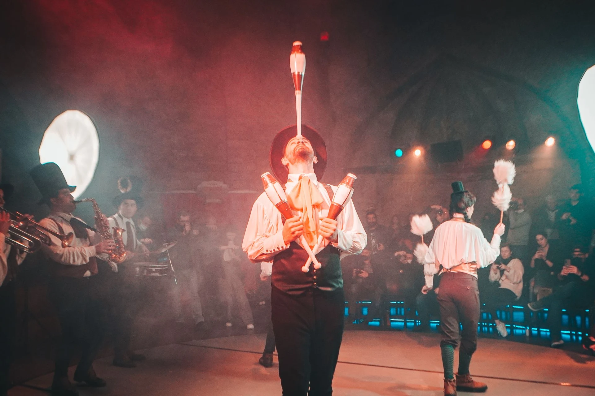 A costumed circus-style performer balancing a club on his chin on a smoky stage, a brass band playing behind him and an audience watching (representative photograph of a variety-theatre performance, not Avanspettacolo Venezia's own cast)